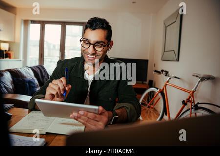 Smiling mixed race businessman interacting on video call holding digital tablet while writing in notepad Stock Photo