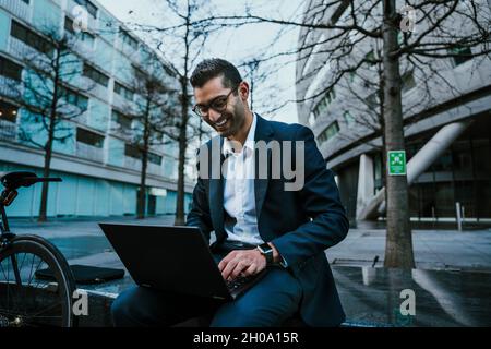 Mixed race businessman typing on laptop smiling while enjoying lunch break  Stock Photo