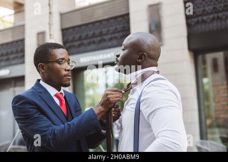 Two black African American businessman friends before an important business meeting. One helps the other to put on a tie and straightens the suit Stock Photo