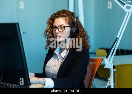 Portrait of a beautiful young woman operator with red curly hair and glasses in the interior of the call center office works at the computer and uses Stock Photo