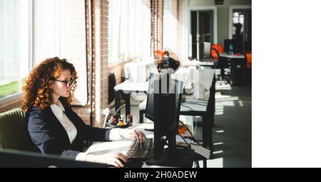 Successful young curly red-haired woman in a business suit and glasses working in an office on a computer Stock Photo