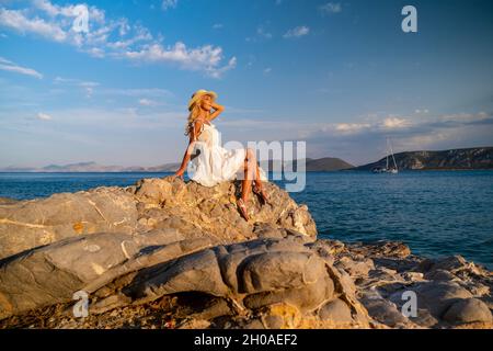 Beautiful young woman wearing traw hat sitting in rocks in white dress by the seashore Stock Photo