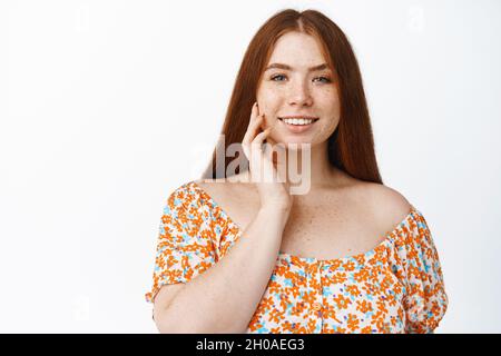 Romantic redhead woman gently touching her face, smiling and gazing carefree at camera, concept of skin and cosmetics, standing over white background Stock Photo