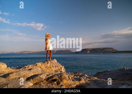 Beautiful young woman wearing traw hat standing in rocks in white dress by the seashore Stock Photo