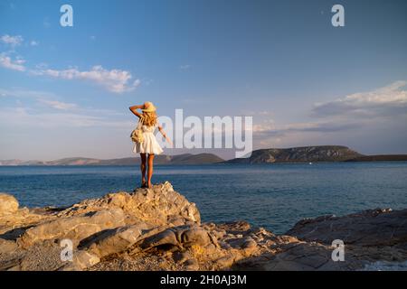 Beautiful young woman wearing traw hat standing in rocks in white dress by the seashore Stock Photo