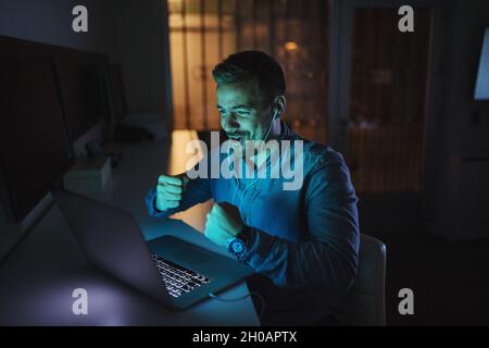 Happy man having video call and gesturing wining while sitting in the office late at night. Stock Photo