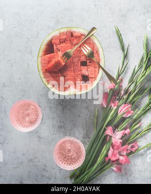 Watermelon cocktails with sparkling wine in wine glasses, sliced watermelon and pink flowers on pale grey concrete kitchen table. Summer celebration w Stock Photo