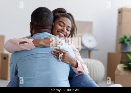 Smiling millennial african american wife hugging husband in new apartment, lady hold keys in room with boxes Stock Photo