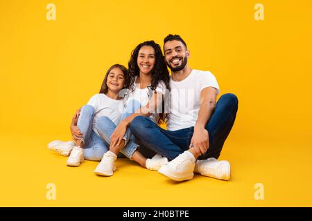 Happy Arabic Father, Mother And Daughter Embracing Sitting, Yellow Background Stock Photo