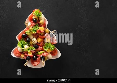 Festive appetizer of canapes, tomatoes, olive, vegetables, mozzarella cheese for festive Xmas party in plate as Christmas tree on black background. Vi Stock Photo
