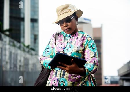 gorgeous african woman walking in the street all merry with a tablet in her hand and wearing a hat on the nature, doing gestures with her hands while Stock Photo