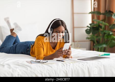 Contented Black Woman Using Phone Wearing Wireless Headphones At Home Stock Photo