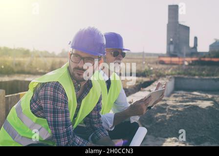 Two engineers with helmets and vests looking at tablet and resting at building site Stock Photo