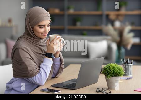 Muslim lady watching video on pc, holding cup drinking coffee Stock Photo