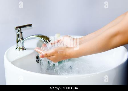 Washing hands in a round white ceramic basin, soap bubbles on hand. There is a green liquid soap bottle on the basin and water is flowing from the tap Stock Photo