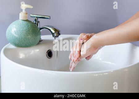 Washing hands in a round white ceramic basin, soap bubbles on hand. There is a green liquid soap bottle on the basin and water is flowing from the tap Stock Photo