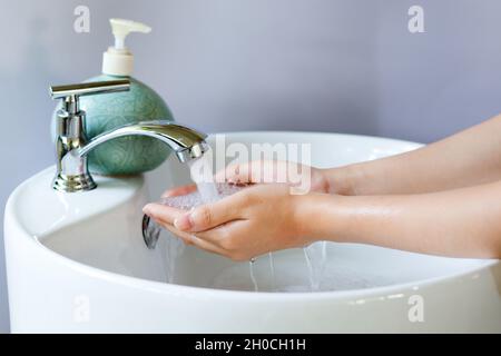 Washing hands in a round white ceramic basin, soap bubbles on hand. There is a green liquid soap bottle on the basin and water is flowing from the tap Stock Photo