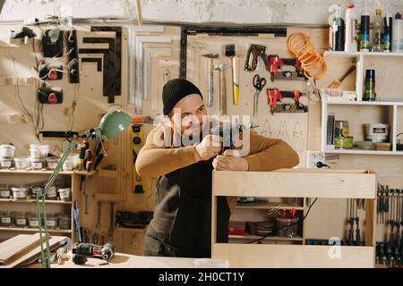 Childish carpenter in a watch cap in a big workshop. Playing with a drill, holding it like a gun and aiming from cover. Number of tools hanging on the Stock Photo