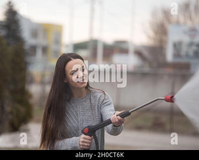 Pretty young woman washing car with manual pressurised water jet in outdoor car-wash Stock Photo