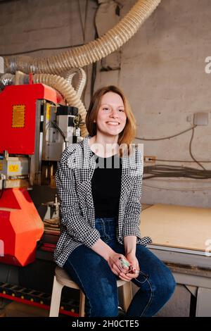 Female worker in semi official clothes sitting on a stool next to a big red piece of heavy machinery Stock Photo
