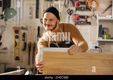 Perky smiling carpenter in a watch cap working in a big workshop. Measuring a piece of wood. Number of tools hanging on the wall. Stock Photo