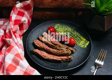 Grilled Picanha steak with chimichurri sauce and cherry tomatoes on a black plate. Fork, checkered napkin on the side. Stock Photo