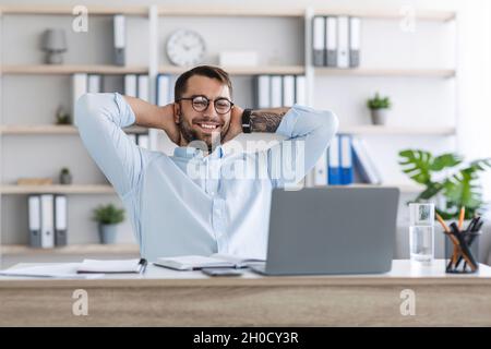 Glad mature european man with beard in glasses with hands behind head resting from remote work behind laptop Stock Photo
