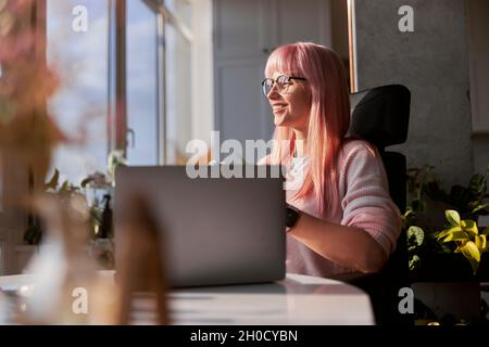 Pink haired woman with elegant glasses works on laptop near window at home Stock Photo