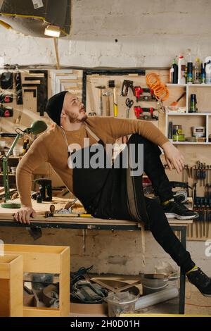 Chilly carpenter in a watch cap sitting on a work table, in a big workshop. Number of tools hanging on the wall. Stock Photo