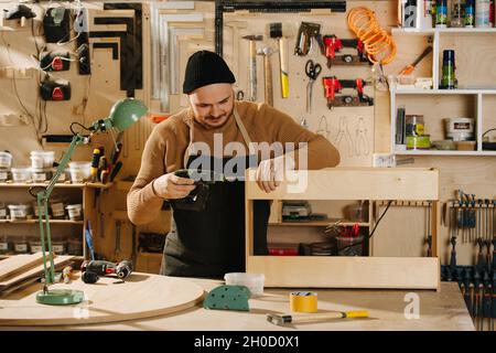 Working carpenter in a watch cap busy in a big workshop. Drilling wooden piece Number of tools hanging on the wall. Stock Photo