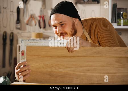 Happy carpenter in a watch cap working in a big workshop. Cheking angle on a piece of wood. Number of tools hanging on the wall. Stock Photo