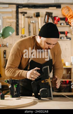 Keen carpenter in a watch cap working in a big workshop. Checking electric plane Number of tools hanging on the wall. Stock Photo