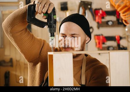Busy carpenter in a watch cap working in a big workshop. Drilling wooden piece Number of tools hanging on the wall. Stock Photo