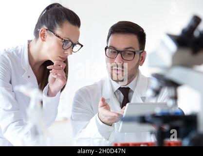 Man and woman scientists looking at tablet and analyzing data from microscope test Stock Photo