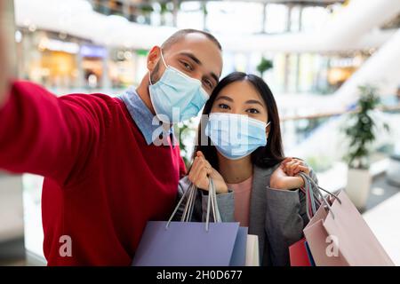 Young multiracial couple in face masks taking selfie together while shopping at huge mall Stock Photo