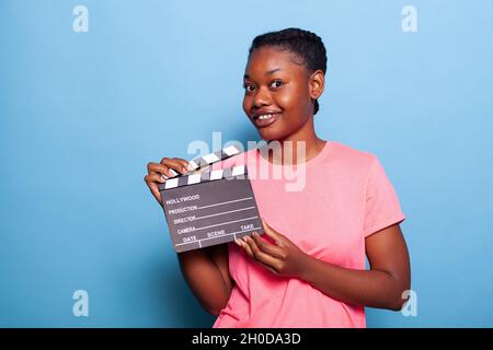 Portrait of smiling african american young woman holding movie production blackboard standing in studio witg blue background. Teenager giving audition for movie roll. Cinematographic concept Stock Photo