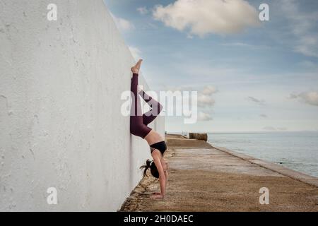 fit woman doing a handstand against a white wall outside by the sea. Stock Photo