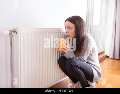 Girl squatting beside radiator with cup of hot tea in hands and trying to warm up Stock Photo