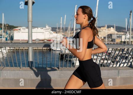 Side view of exhausted young sportswoman in activewear breathing heavily and running on embankment near sea water and sailboats in morning in city. Stock Photo