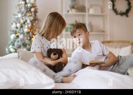 Brother is reading a book to his sister, sitting on the bed. Stock Photo