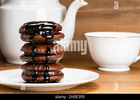 Closeup view stack of delicious choco pie topped with chocolate syrup in white ceramic dish with a cup of hot tea and white kettle on wooden table. Stock Photo