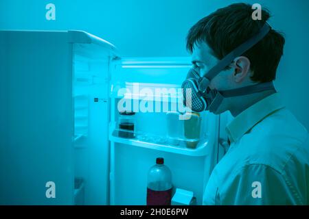 A man wearing an air filtration mask opens a refridgerator door. Perhaps he is concerned about what he may find inside. Stock Photo