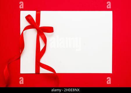 Gift card white blank with red ribbon bow on red background, Empty note paper, copy space, message template. Christmas or valentine present satin curl Stock Photo