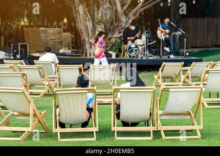 Back view of Asian young couple sit to relax on white deck chairs with tables for dinner in lawn is surrounded by shady green grass with blurred image Stock Photo