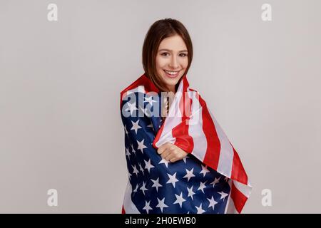 Portrait of friendly young woman standing wrapped in American flag looking at camera and smiling, wearing white casual style T-shirt. Indoor studio shot isolated on gray background. Stock Photo