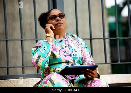 charming african woman sitting on the stairs at the back of the office, holding a tablet in her hand and talking while she was talking on the phone. Stock Photo