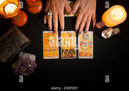 Detail of a woman's hands showing three tarot cards face up, on a black card table. Concept of divination session with tarot cards. View from above. Stock Photo