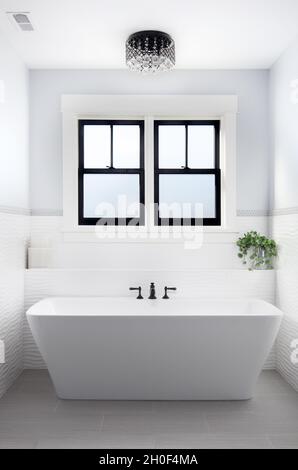 A beautiful, luxury bathroom with a light hanging above a white Fleurco standalone tub and black faucet surrounded by tiles. Stock Photo