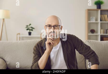 Webcam view of serious adult man sitting on sofa at home, looking at camera and thinking Stock Photo
