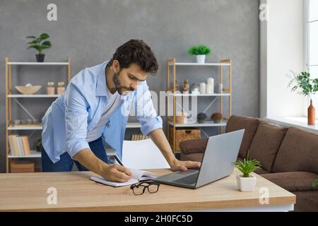 Man standing by desk, using laptop computer and writing down information in notebook Stock Photo